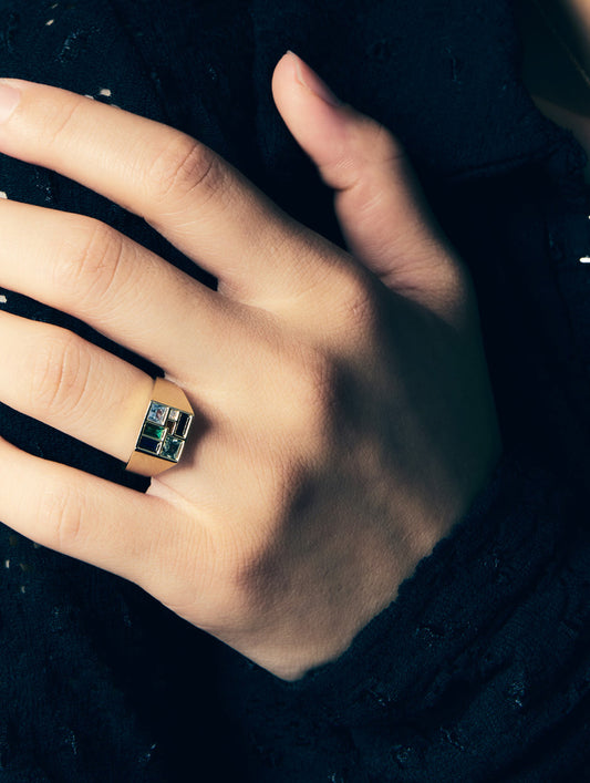 A close-up of a hand wearing a gold signet ring with a rectangular green and blue inlay, set against a dark textured background.