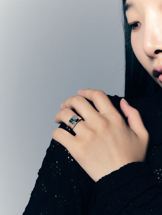 A close-up lifestyle shot of a model wearing a silver ring with a rectangular blue and green stone, set against a plain gray background.