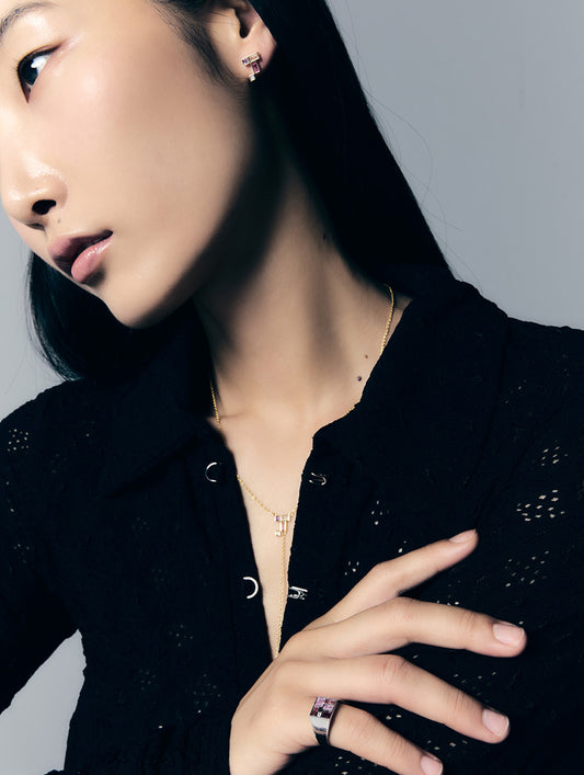 A close-up lifestyle shot of a model wearing a gold geometric pendant necklace and a matching gold ring with a purple gemstone, styled with a black textured top against a neutral gray background.