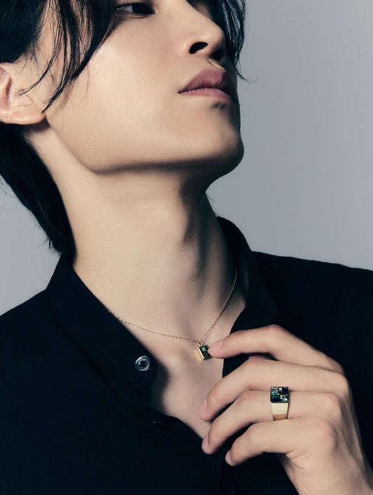 A close-up lifestyle shot of a model wearing a delicate gold chain necklace with a small rectangular pendant and a gold signet ring with a black and white patterned face, paired with a black collared shirt against a neutral gray background.