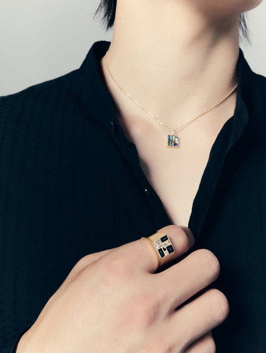 A close-up lifestyle shot of a person wearing a delicate gold chain necklace with a small square pendant featuring blue and green inlay, paired with a black ribbed shirt and a matching gold ring on their finger.