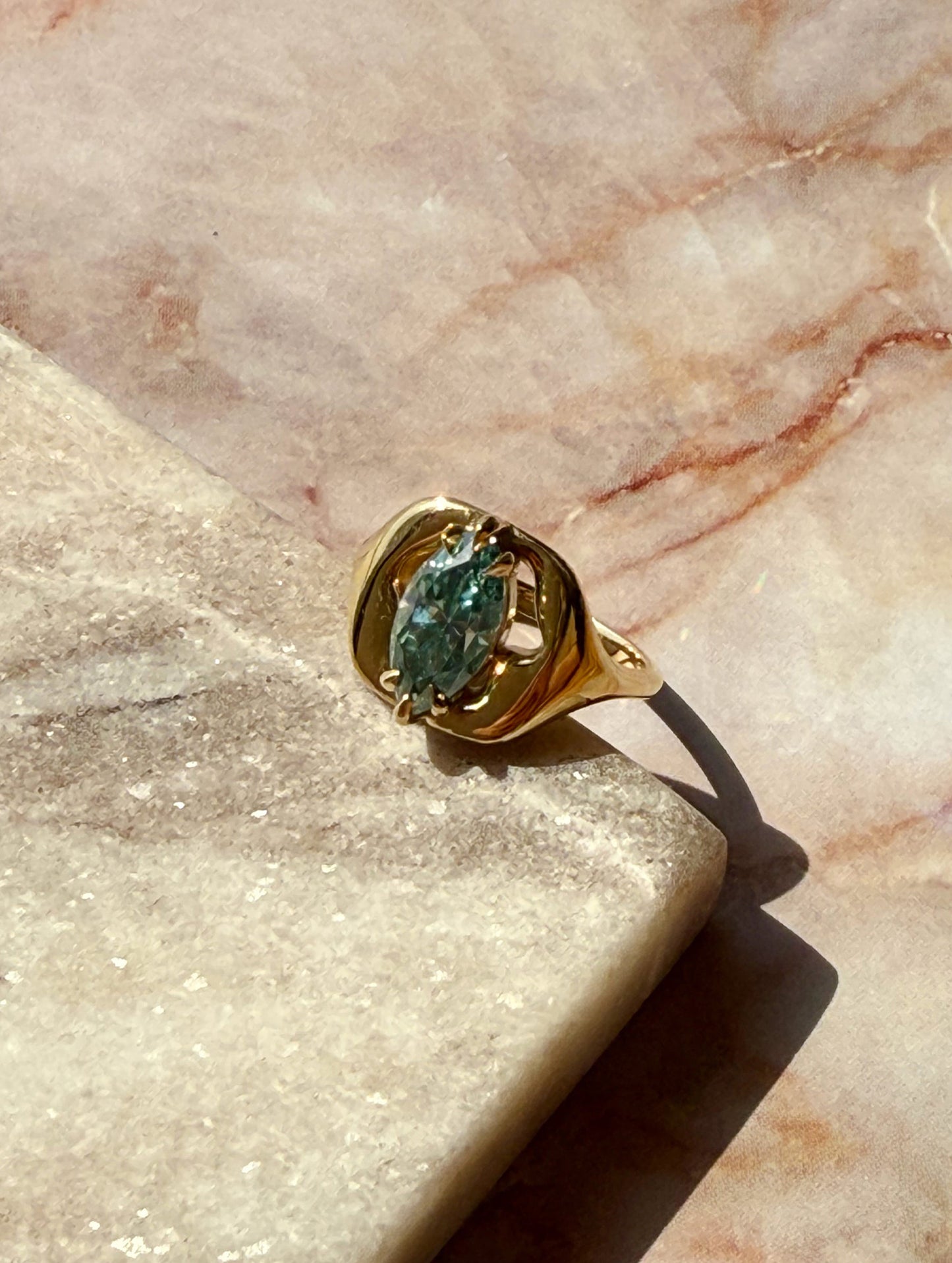 A gold ring with a marquise-cut green gemstone, resting on a textured beige stone slab against a pinkish marble background.