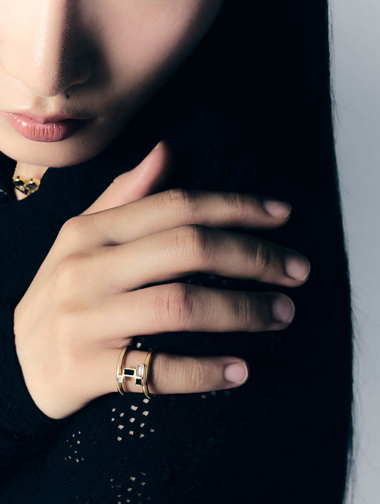 A close-up lifestyle shot of a model's hand wearing two gold rings with black rectangular inlays, set against a dark background.