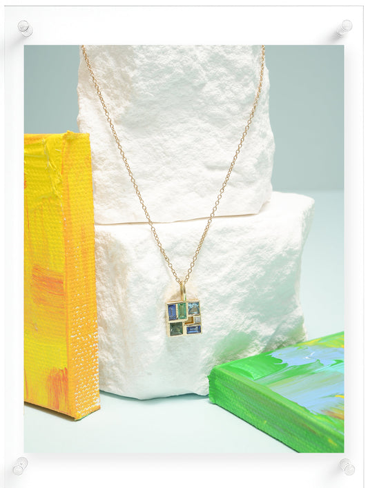 A gold necklace with a geometric mosaic pendant featuring blue, green, and yellow inlay, displayed on a white textured stone block with abstract colorful art canvases in the background.