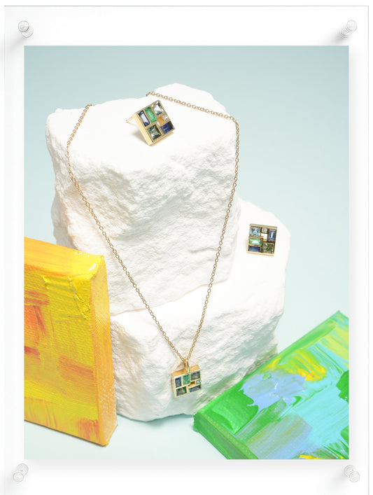 A gold chain necklace and matching earrings with square colorful enamel pendants, displayed on a white textured stone block with abstract painted blocks in the background.