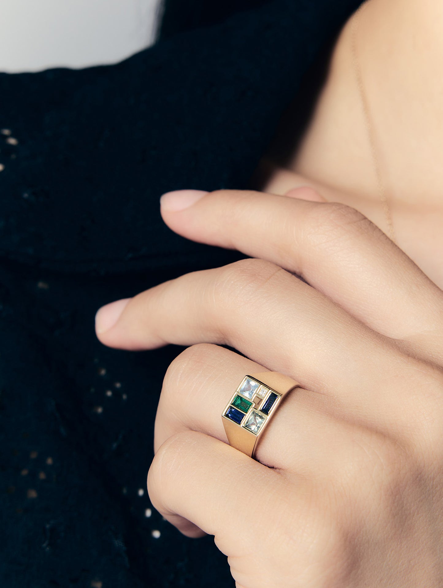 A close-up of a person's hand wearing a gold signet ring with a geometric, multi-colored face featuring green, blue, and clear stones, set against a dark textured garment.