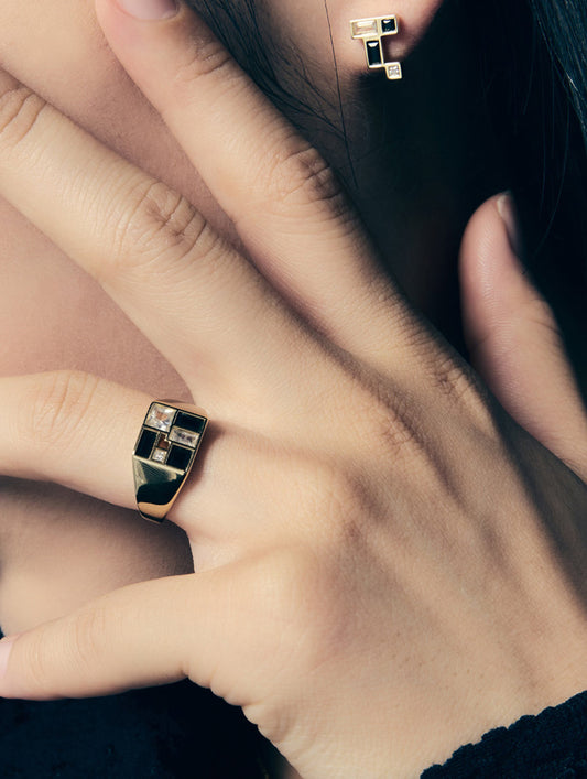 A close-up lifestyle shot of a person's hand wearing a gold signet ring with a geometric black and white patterned face, with a matching geometric stud earring visible on the ear.
