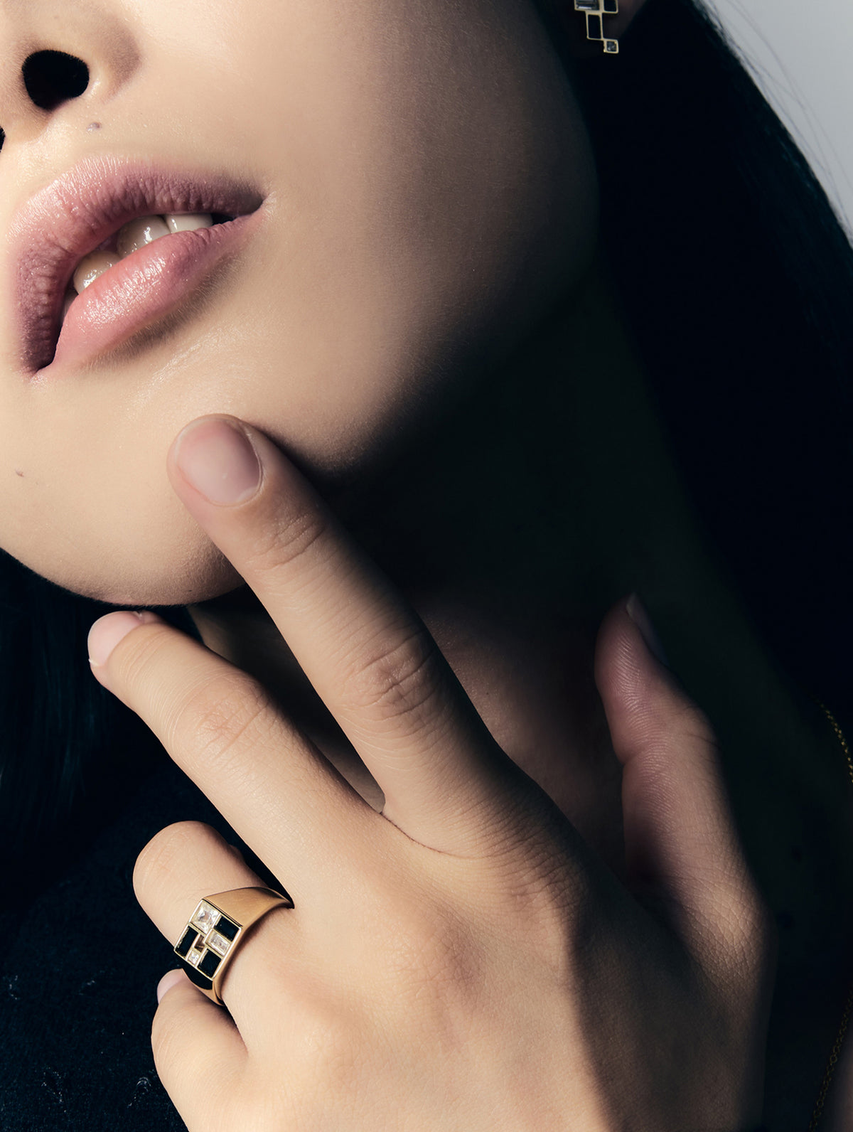 Close-up of a person's hand and lower face wearing a gold ring with a black geometric block design, against a dark background.