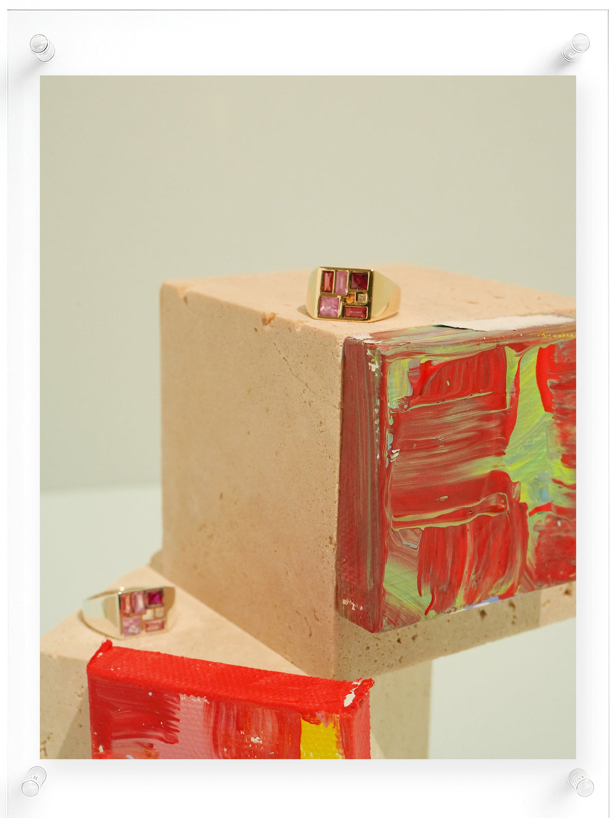 Two gold rings with square pink and red gemstone settings displayed on beige concrete blocks with red and green painted textures, set against a light beige background.