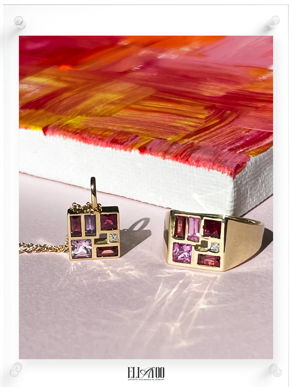 Gold jewelry set featuring a square pendant necklace and matching ring with rectangular magenta and purple gemstones, displayed on a light pink surface in front of a colorful abstract canvas.