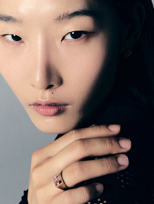 A close-up portrait of a model wearing a gold ring with pink gemstones on her finger, set against a dark background.