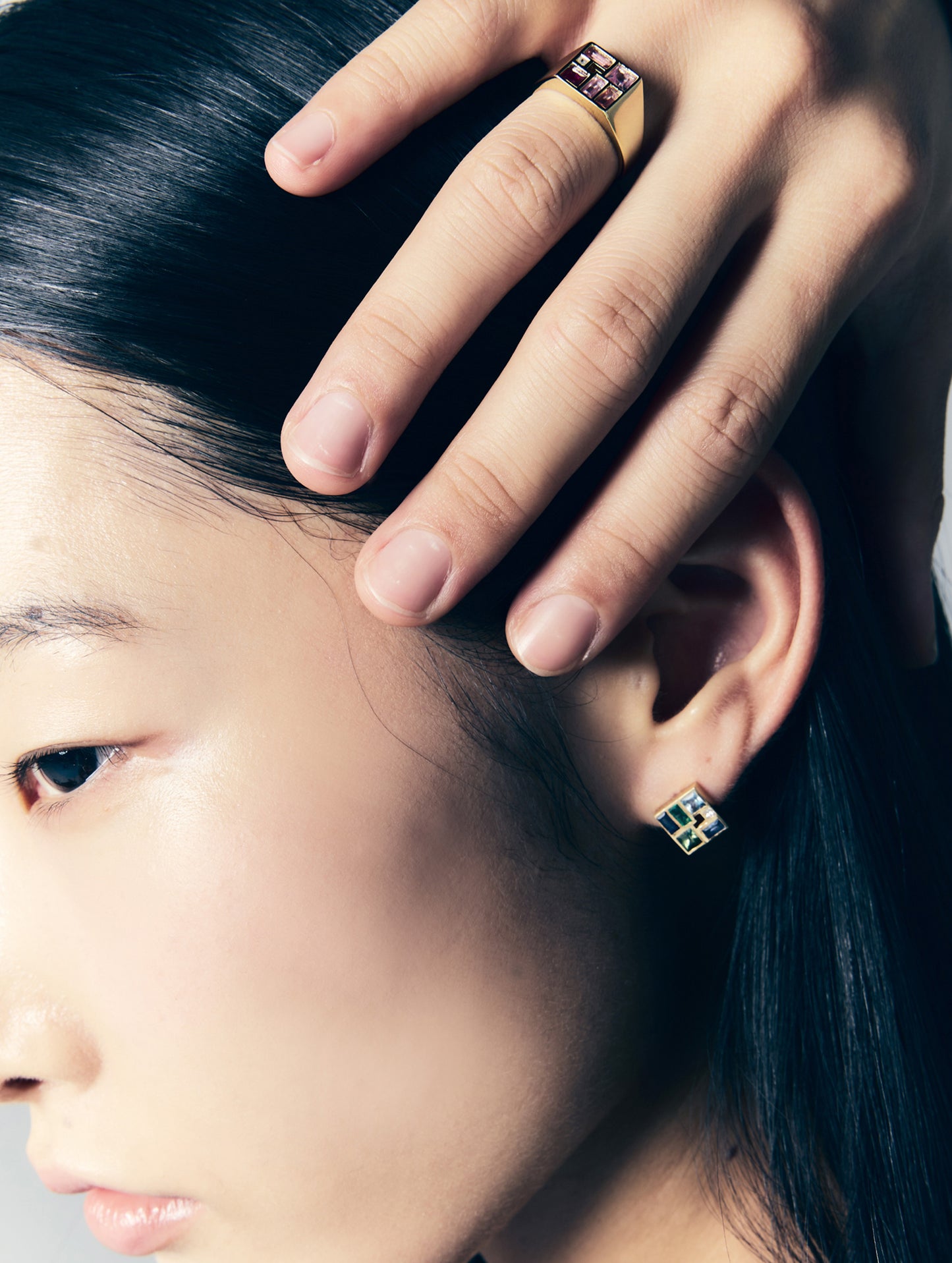 Close-up of a model wearing a gold signet ring with a purple and green stone, along with matching green and blue stud earrings.