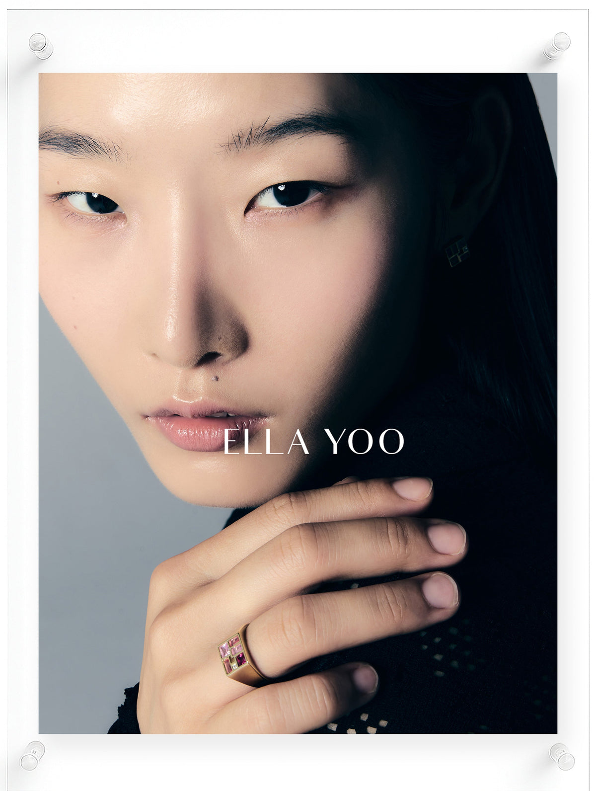 A close-up portrait of a model wearing a gold ring with a pink gemstone, set against a dark background with the brand name 'ELLA YOO' printed across the center.