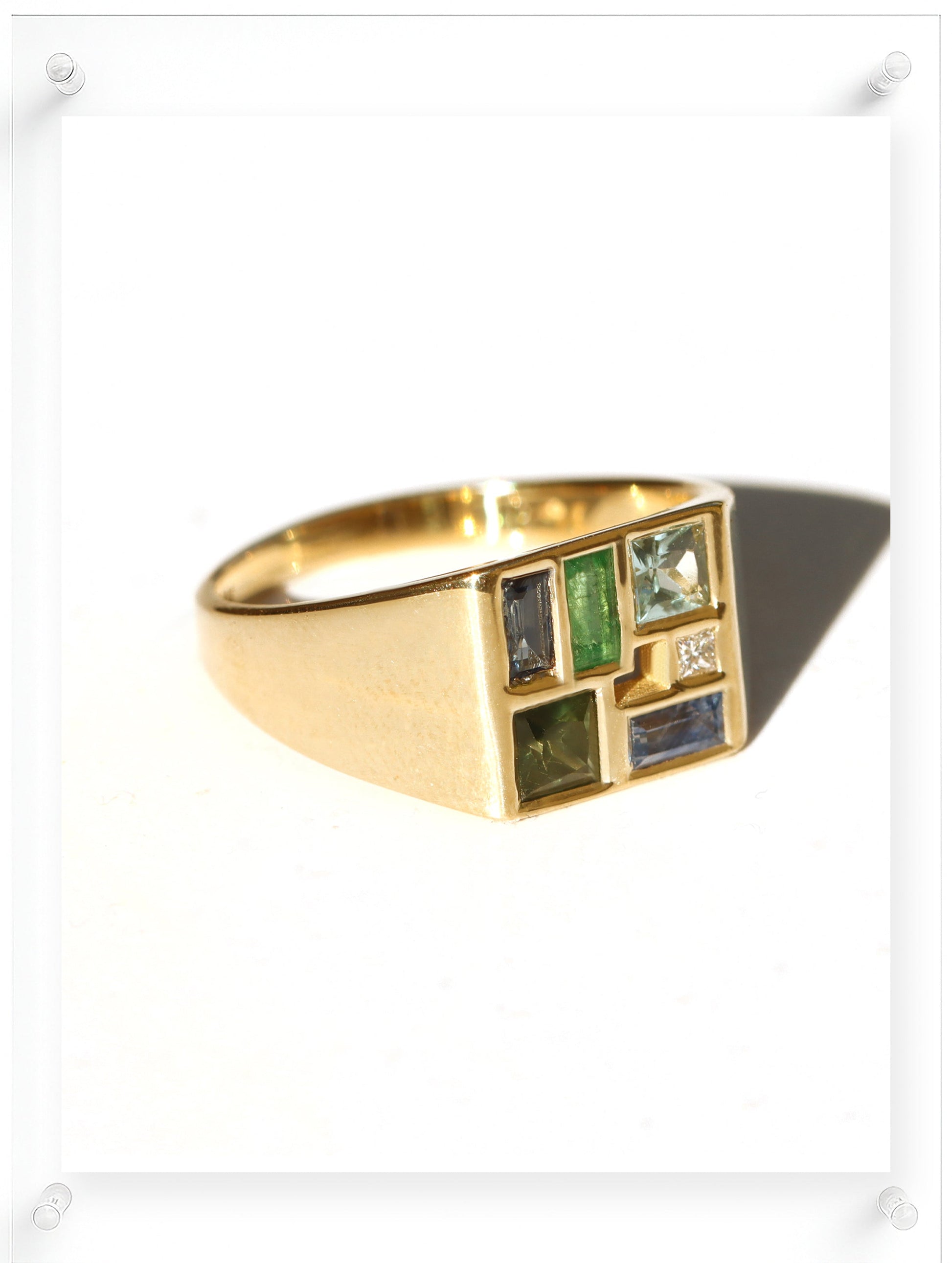 A gold signet ring with a geometric face featuring multiple inset rectangular gemstones in green, blue, and clear, displayed on a clean white background.
