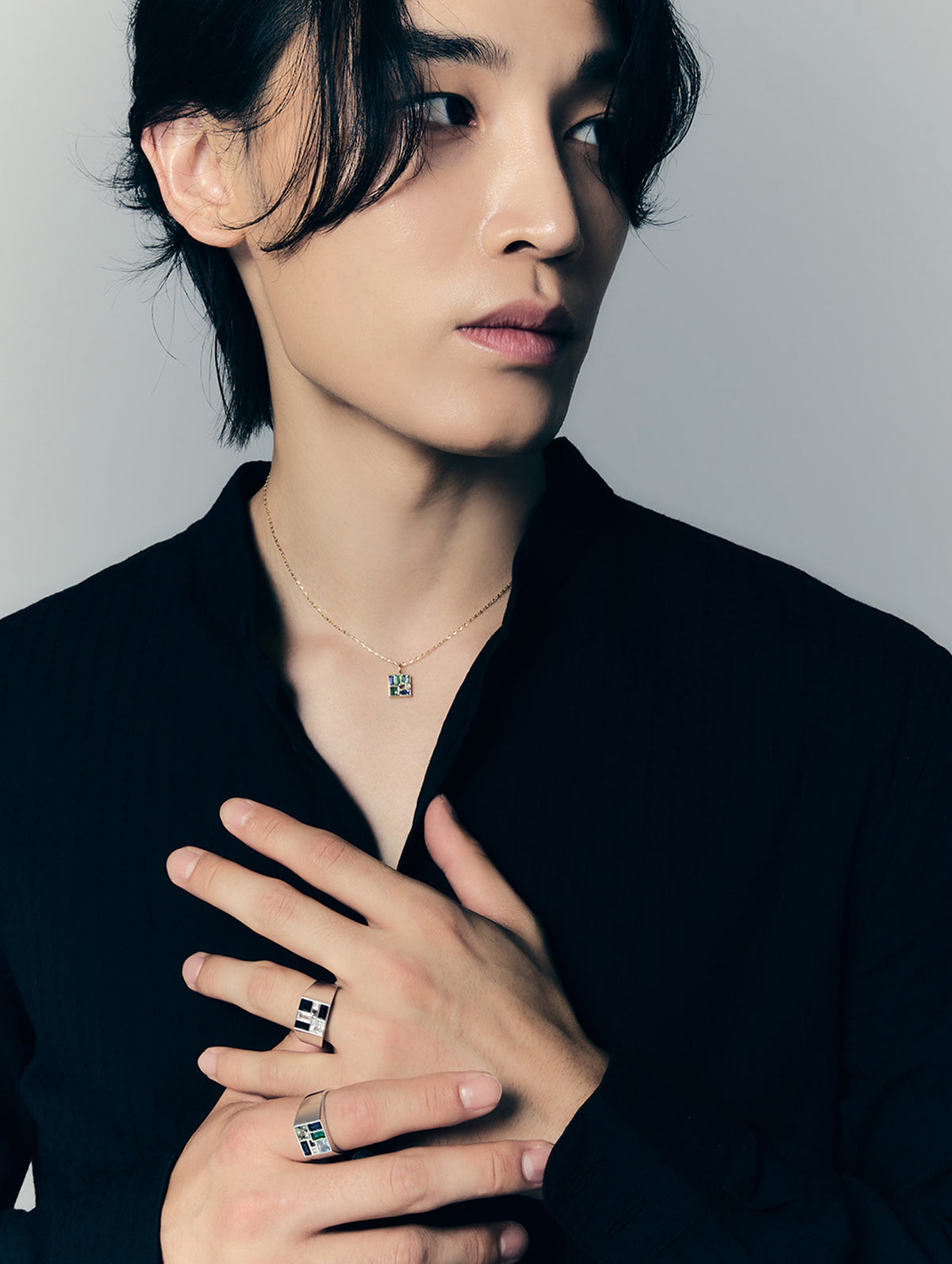 A close-up portrait of a man wearing silver geometric rings on his fingers and a delicate necklace, dressed in a black shirt against a neutral gray background.
