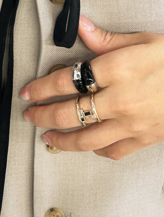 A close-up of a hand wearing three rings: a wide black ring, a slim silver ring, and a gold ring with a rectangular black inlay, set against a beige tweed jacket.