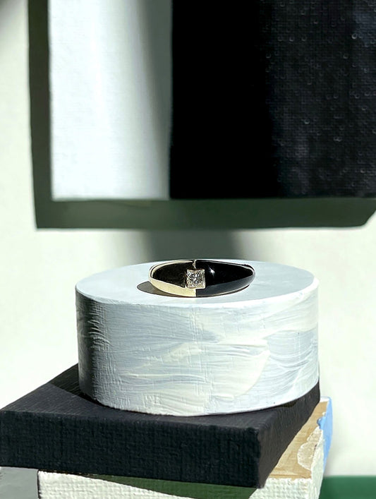 A gold ring with a clear gemstone set in a black band, displayed on a cylindrical white textured pedestal atop stacked black and beige blocks.