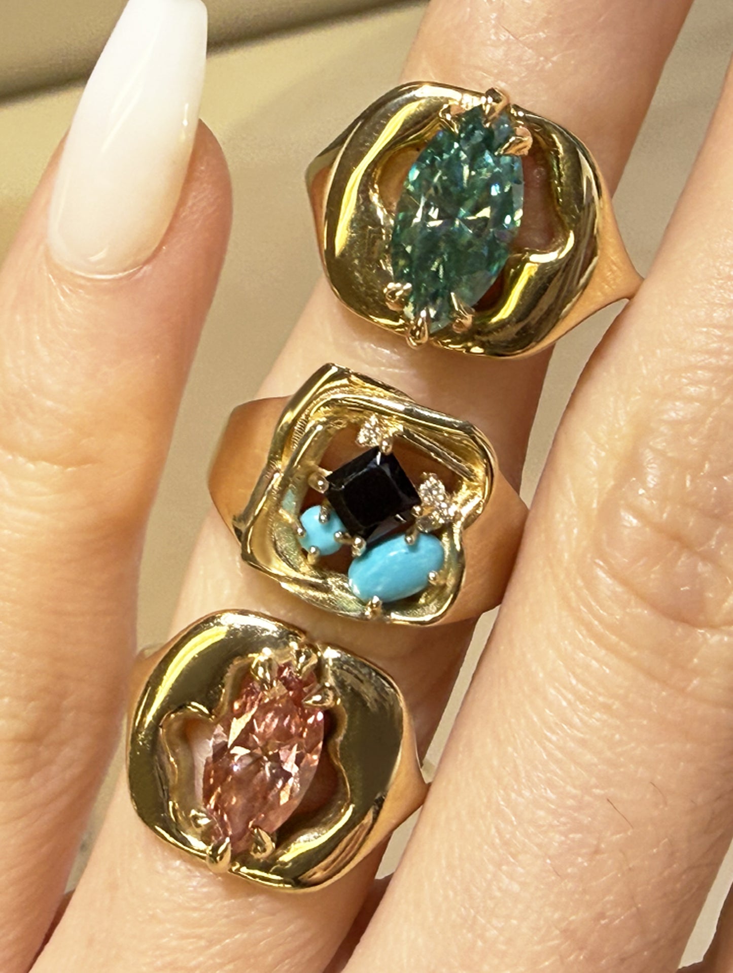 Three gold statement rings with different colored stones are shown worn on a hand. The top ring features a marquise-cut teal stone, the middle ring has a square black stone with turquoise and small clear stones, and the bottom ring has a marquise-cut pink stone.