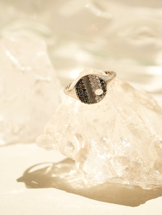 A silver signet ring with a black and white yin-yang symbol face, resting on a textured clear crystal block against a warm, sunlit beige background.